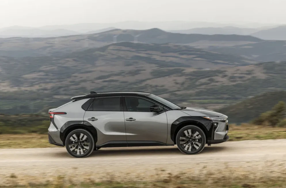 Sivi krosover automobil u pokretu, sa crnim detaljima i felgama, vozi se po makadamskom putu sa brežuljcima i planinama u pozadini.