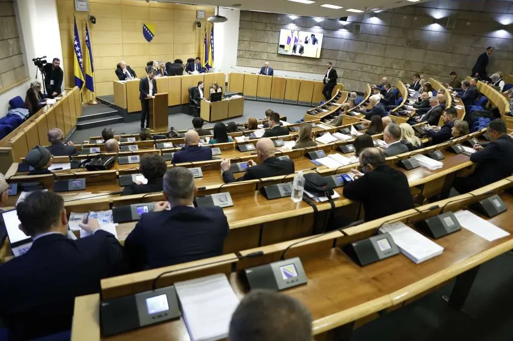 Dugačak ambijent parlamentarne dvorane sa drvenim klupama okrenutim prema podijumu. Nekoliko ljudi sjedi na klupama, okrenutih leđima prema kameri, a fokus je na govorniku koji stoji na podijumu.