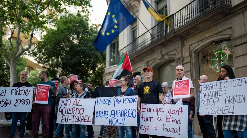 Grupa ljudi na demonstracijama ispred zgrade sa zastavama EU i Palestine, drže transparente sa natpisima 'NO BUSINESS AS USUAL WITH ISRAEL', 'NO AL ACUERDO EU-ISRAEL', 'ARMS EMBARGO NOW!', 'ISRAELI CITIZEN DEMAND: STOP ARMING ISRAEL!' i 'Europe, don't turn a blind eye to Gaza!'