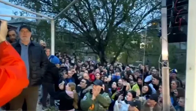 Skupina ljudi na otvorenom, fokus na muškarca u crnoj jakni i plavom džemperu.