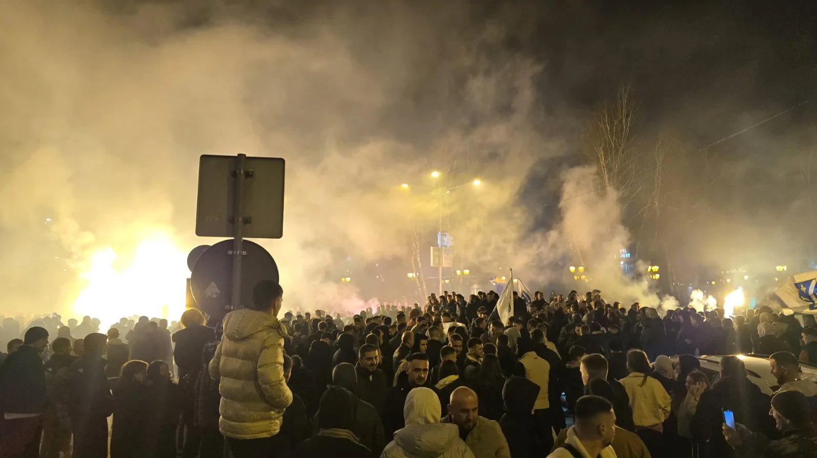 Velika grupa ljudi, navijača, okupljena na ulici noću, uz mnogo dima od baklji i osvijetljene gradske lampe u pozadini.