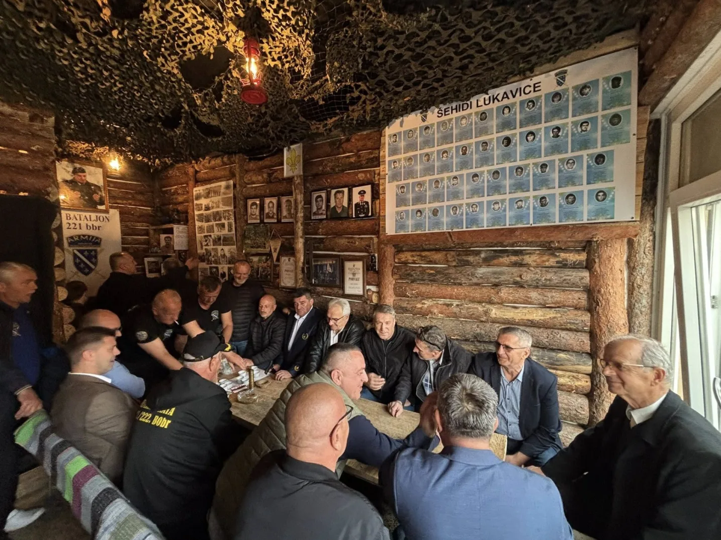 Skupina muškaraca, vjerojatno ratnih veterana, okupljena u drvenoj prostoriji. Na zidovima su slike i pano s fotografijama poginulih boraca s natpisom "ŠEHIDI LUKAVICE".