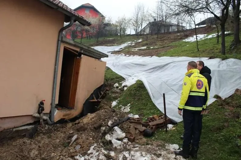 Dvije osobe u zaštitnoj opremi promatraju oštećenu kuću i klizište prekriveno bijelom ceradom.