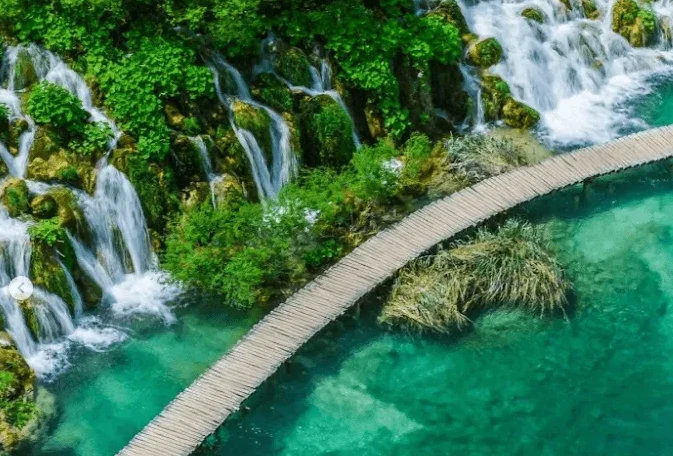 Drveni most vijugavog oblika koji premošćuje tirkiznu vodu pored brojnih slapova okruženih bujnim zelenilom.