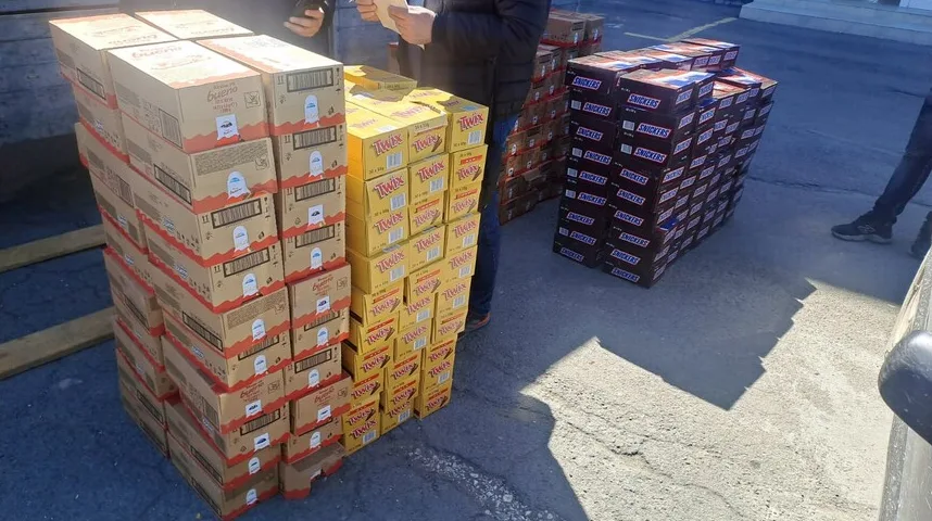 Velike hrpe kutija sa slatkišima, uključujući Kinder Bueno, Twix i Snickers, posložene na paletama na otvorenom.