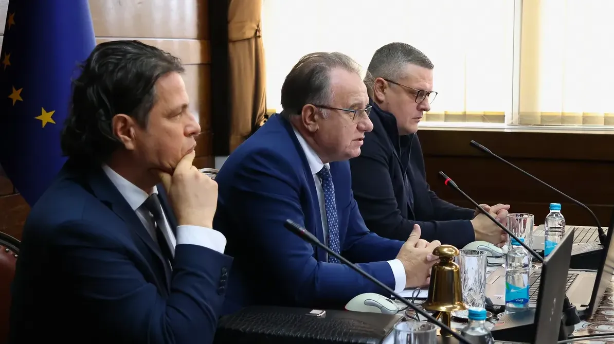 Tri muškarca sjede za stolom na sastanku, jedan drži ruku na bradi, drugi govori, treći gleda ispred sebe.