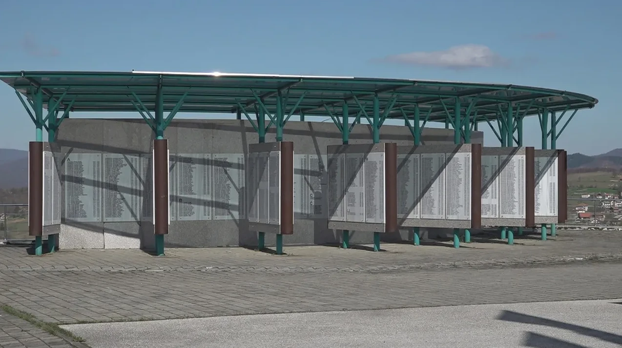 Moderni spomenik sa zelenom nadstrešnicom i stupovima, na zidovima su ispisana imena u više kolona, te pogled na naselje u pozadini.