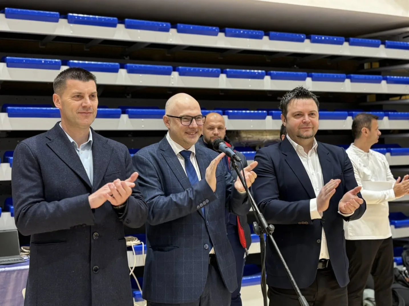 Tri muškarca u odijelima aplaudiraju na podijumu ispred tribina plave boje.