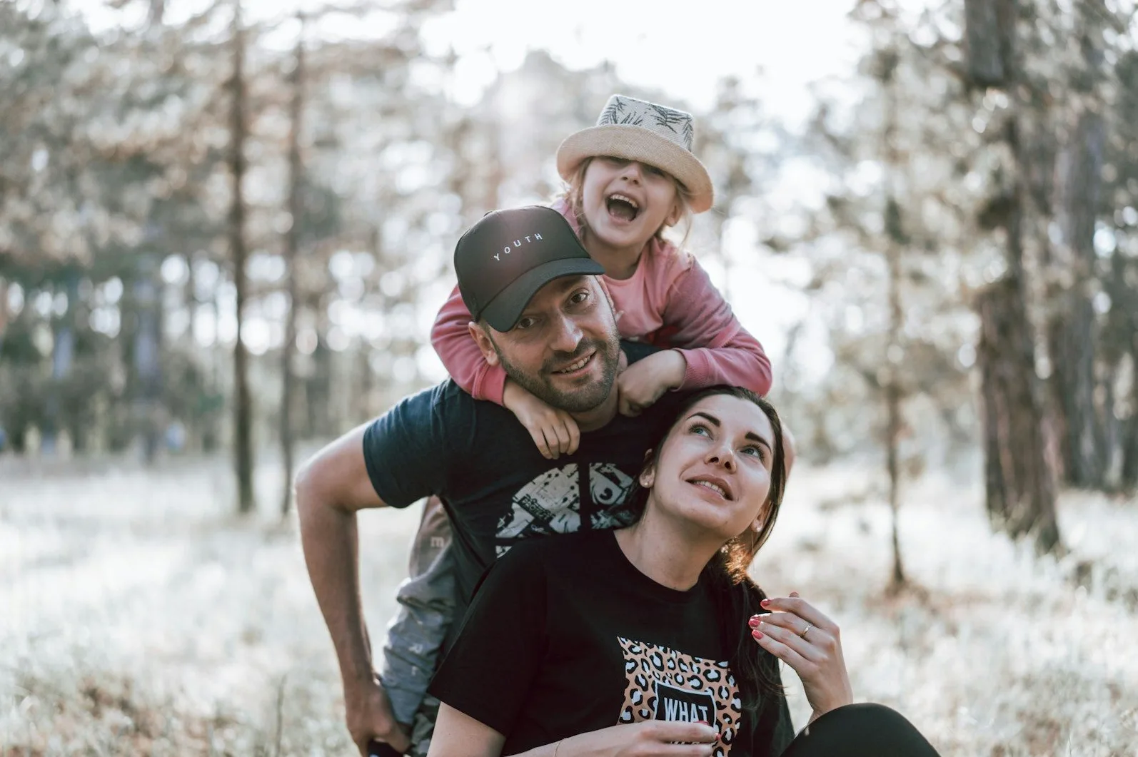 Otac nosi kćerku na ramenima dok majka sjedi pored njih u prirodi.