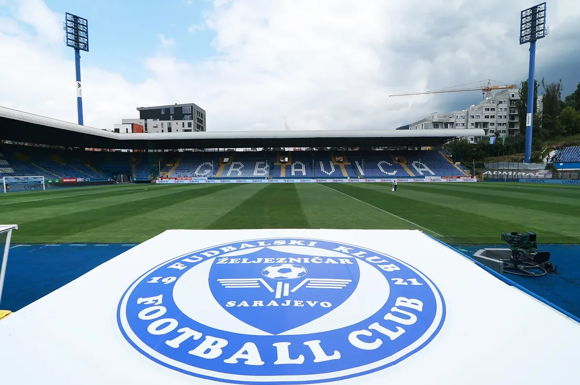 Stadion Grbavica u Sarajevu, dom FK Željezničar, sa istaknutim grbom kluba u prvom planu i tribinama koje ispisuju "GRBAVICA" na bosanskom jeziku.