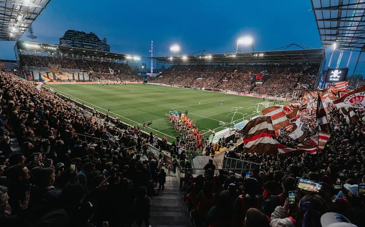 Fudbalska utakmica sa punim stadionom navijača u sumrak, sa svjetlima reflektora koji osvjetljavaju teren.