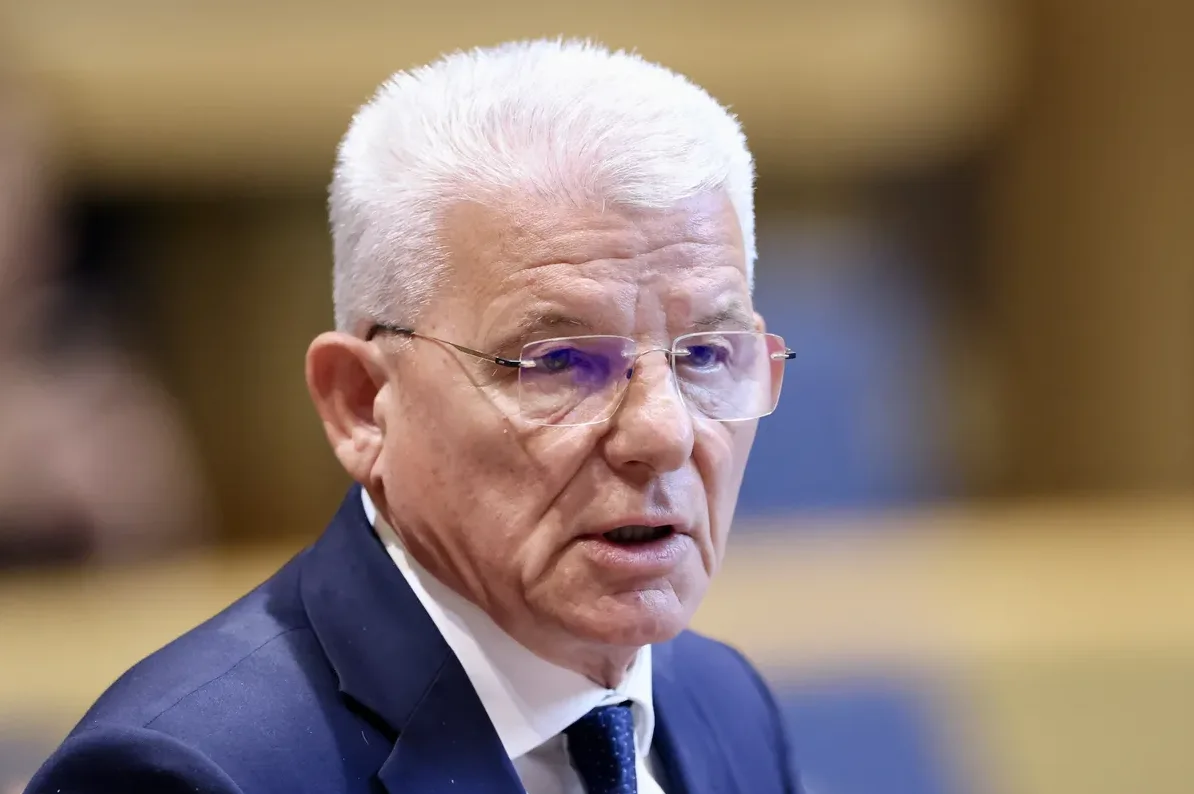 Portret starijeg muškarca bijele kose i naočara, obučenog u tamnoplavo odijelo.