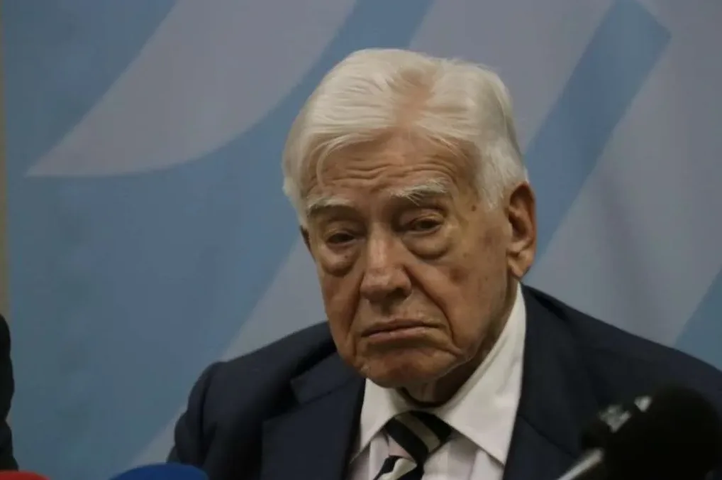 Portret starijeg gospodina sijede kose u tamnom odijelu i bijeloj košulji sa prugastom kravatom, gleda u stranu sa ozbiljnim izrazom lica.