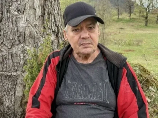 Portret starijeg muškarca sa kapom na glavi, obučenog u crveno-crnu jaknu i majicu sa natpisom "Everlast", kako sjedi pored drveta u prirodi.