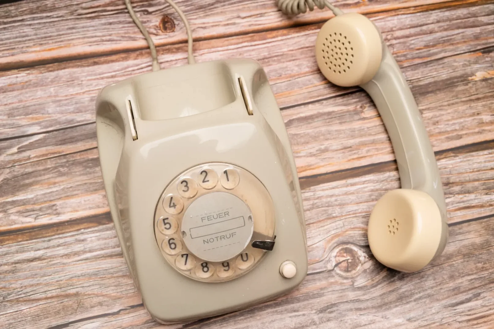 Starinski bež telefon sa kružnim brojčanikom i slušalicom odloženom pored njega, postavljen na drvenu površinu.