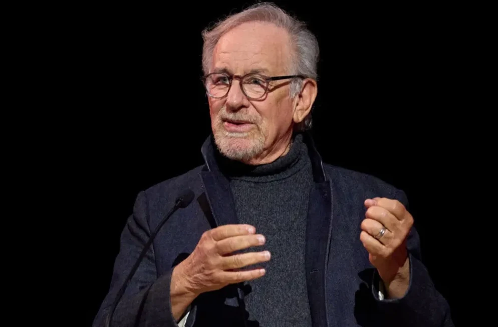 Steven Spielberg u tamnoj dolčeviti i jakni, nosi naočale i govori za govornicom.