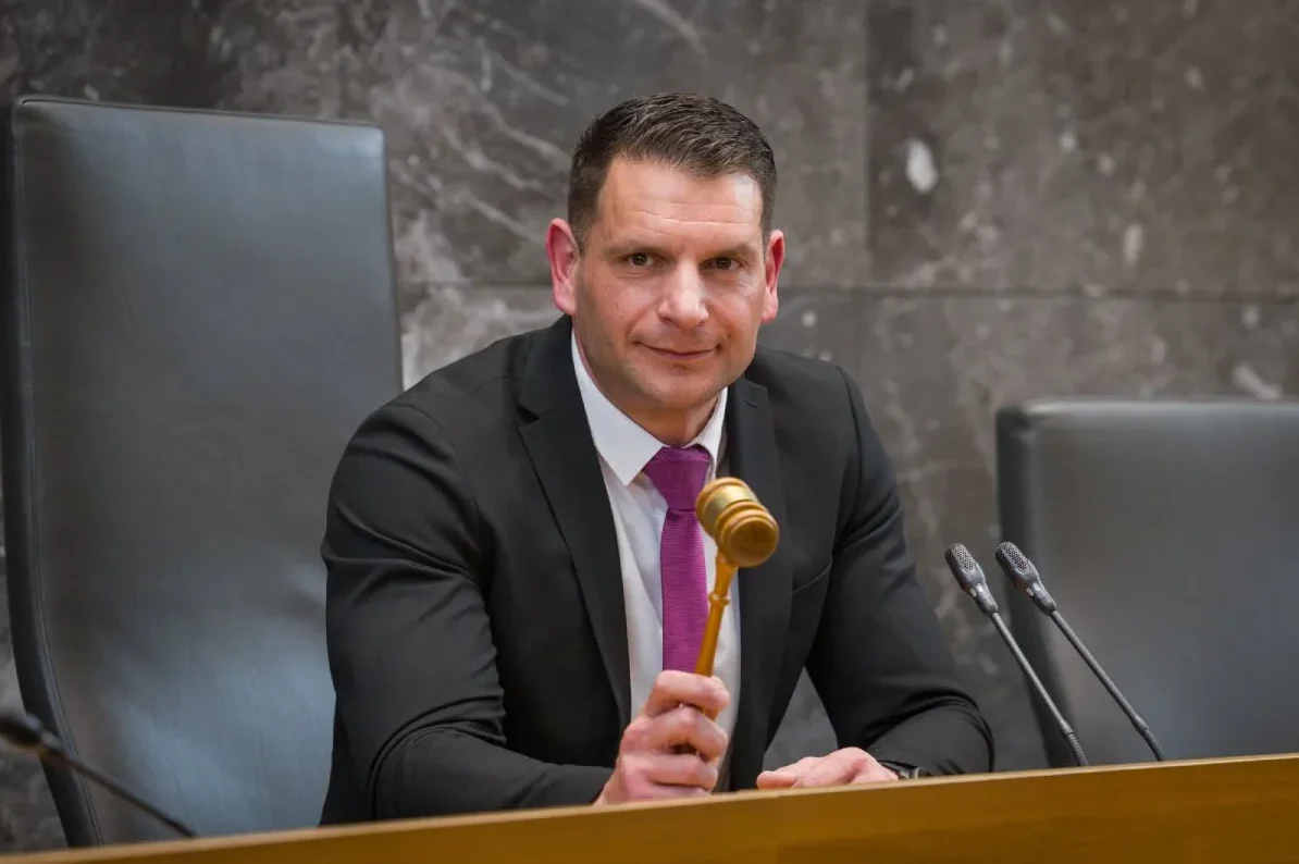 Čovjek u crnom odijelu sa ljubičastom kravatom drži sudijski čekić.