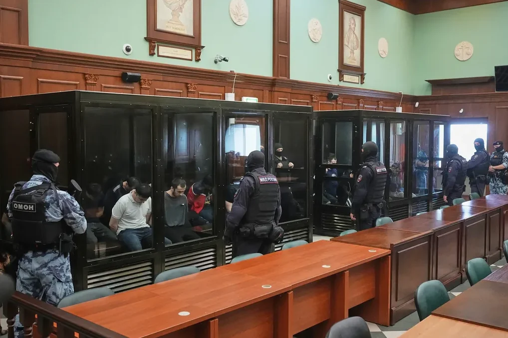 Optuženi sjede u staklenim boksovima u sudnici, dok ih obezbjeđuje naoružana policija u uniformama.