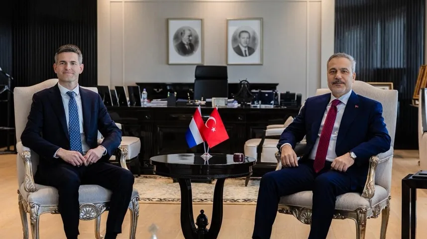 Dva muškarca u odijelima sjede u luksuznim foteljama, dok se između njih na stolu nalaze zastave Holandije i Turske, te portreti na zidu u pozadini.