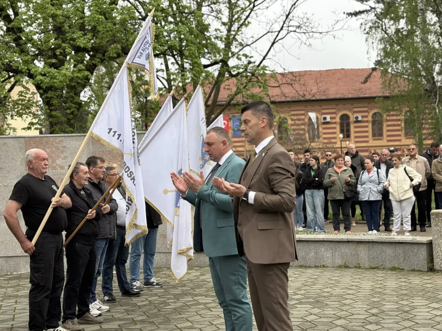 Ljudi u svečanim odijelima drže zastave i polažu ruke u molitvi.