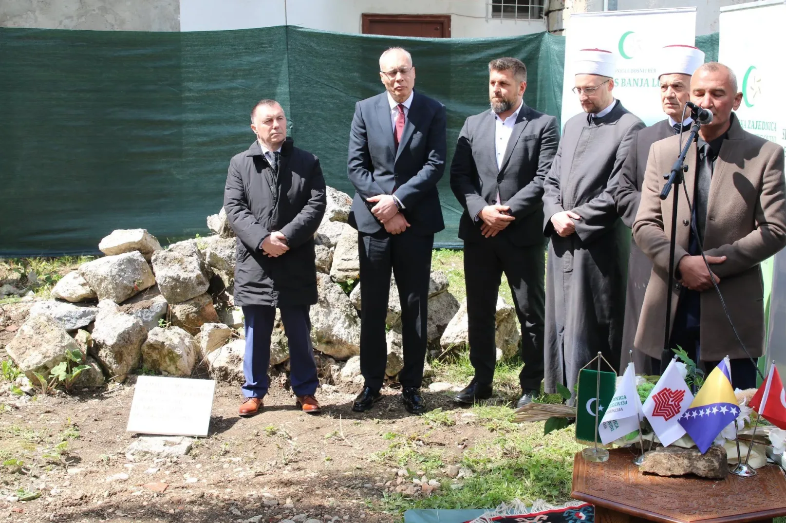 Grupa muškaraca u svečanim odijelima i vjerskim odjevnim predmetima stoji ispred ruševina i zastava.