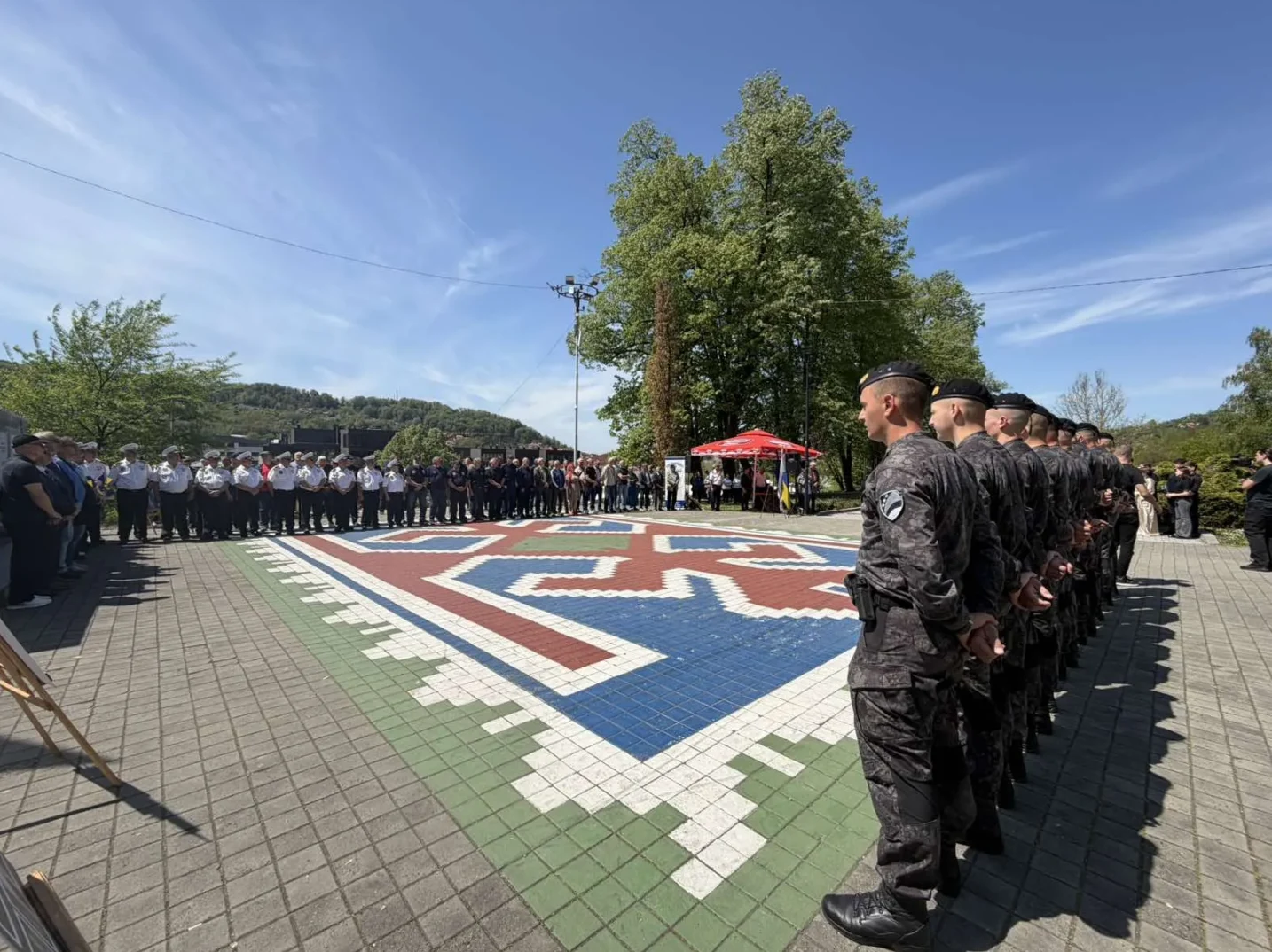 Pukovnija uniformiranih osoba, među kojima su vojnici u tamnim uniformama i civili u bijelim košuljama, stoji postrojena ispred velikog šarenog tepiha postavljenog na popločanom trgu.