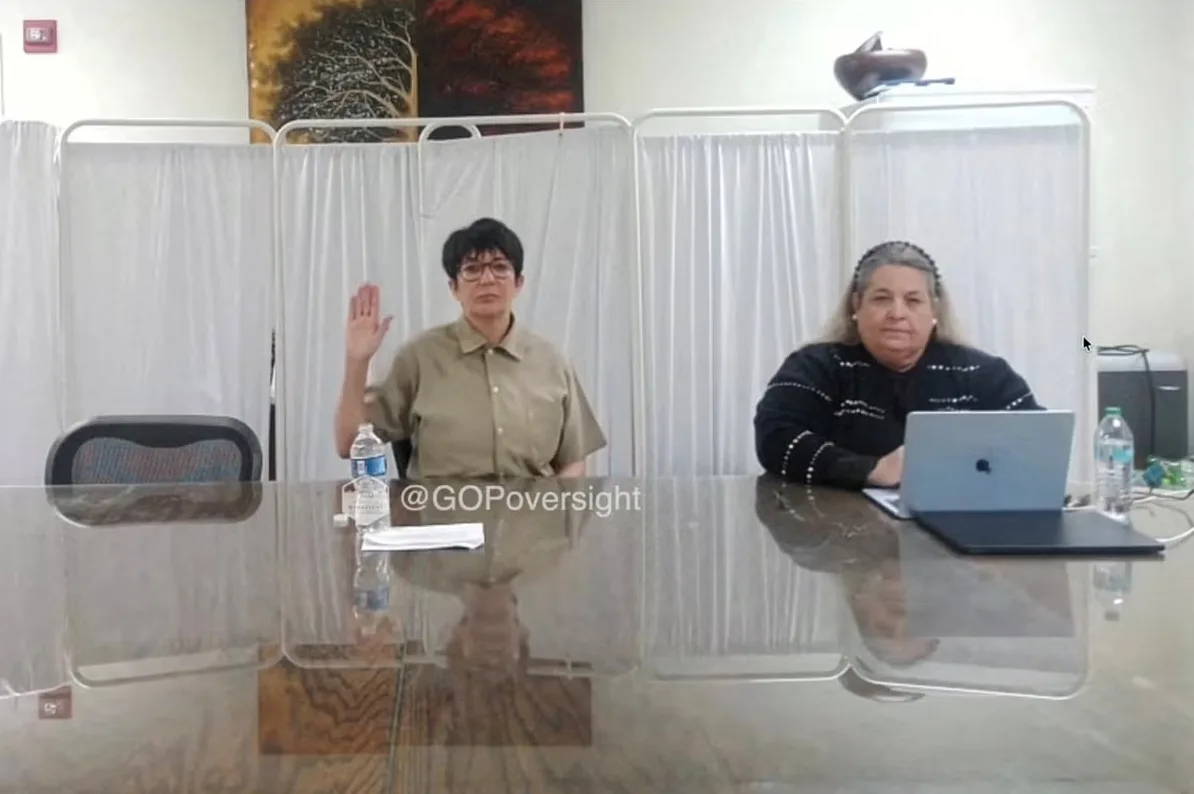 Žena podiže ruku u zrak dok polaže zakletvu, sjedeći za stolom ispred prozora i paravana.