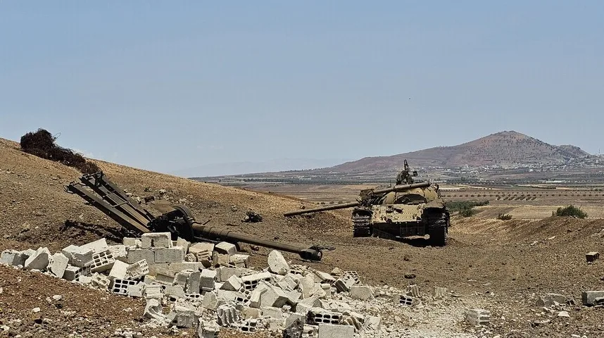 Oštećeni tenk i topovska cijev na brdovitom terenu sa razbacanim betonskim ciglama u prednjem planu i selom u daljini.