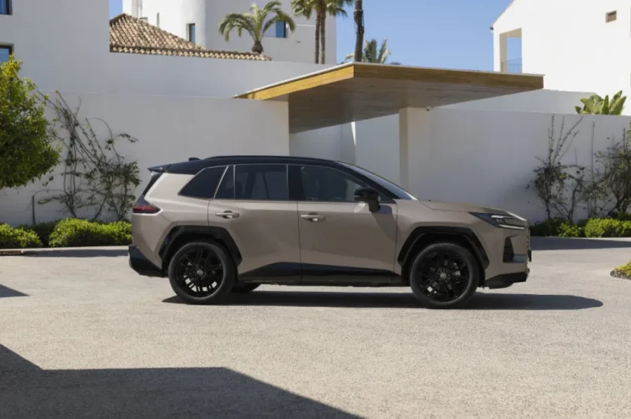 Smeđe terensko vozilo sa crnim krovom i crnim felgama parkirano ispred moderne bijele građevine sa drvenim detaljima i palminim drvećem.