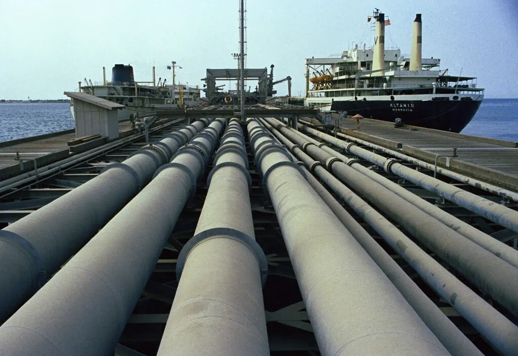 Detaljan pogled na niz velikih sivih naftovoda postavljenih na metalnoj konstrukciji, koji vode prema obali. U pozadini se vidi veliki teretni brod nazvan 'ALTANIN' koji je usidren uz pristan. Nebo je vedro i plavo, a more mirno.