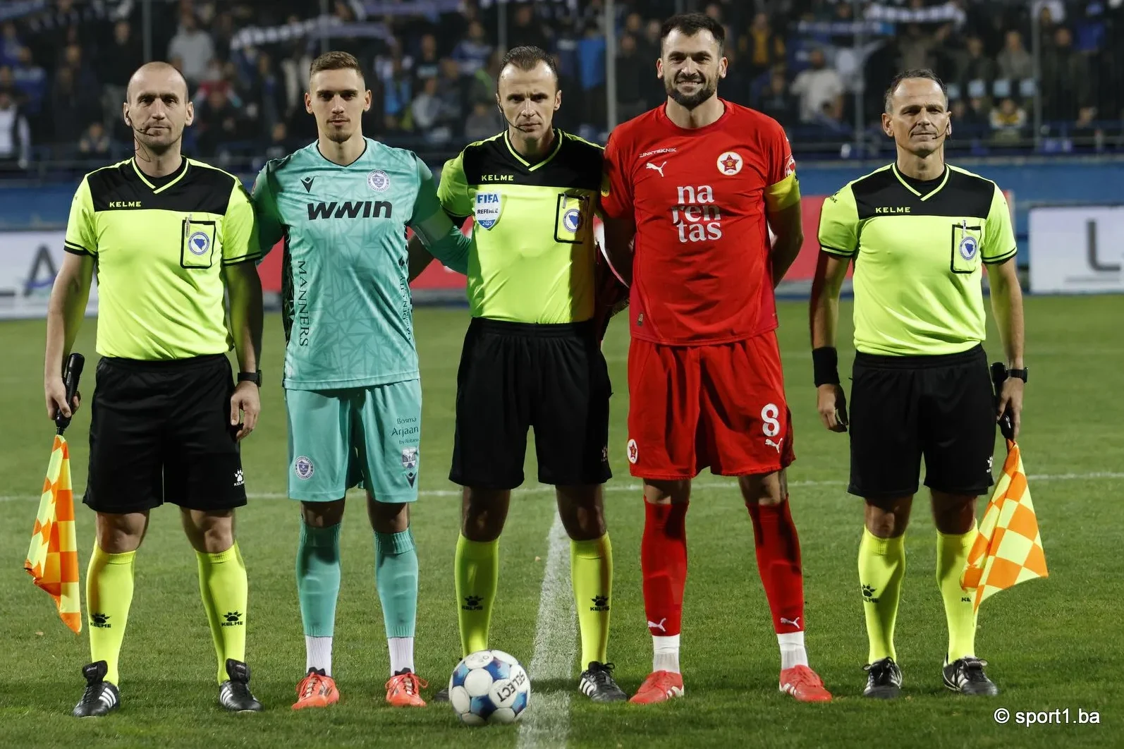 Fudbalski tim u crvenim dresovima i sudije u zelenim majicama stoje na terenu pred utakmicu.