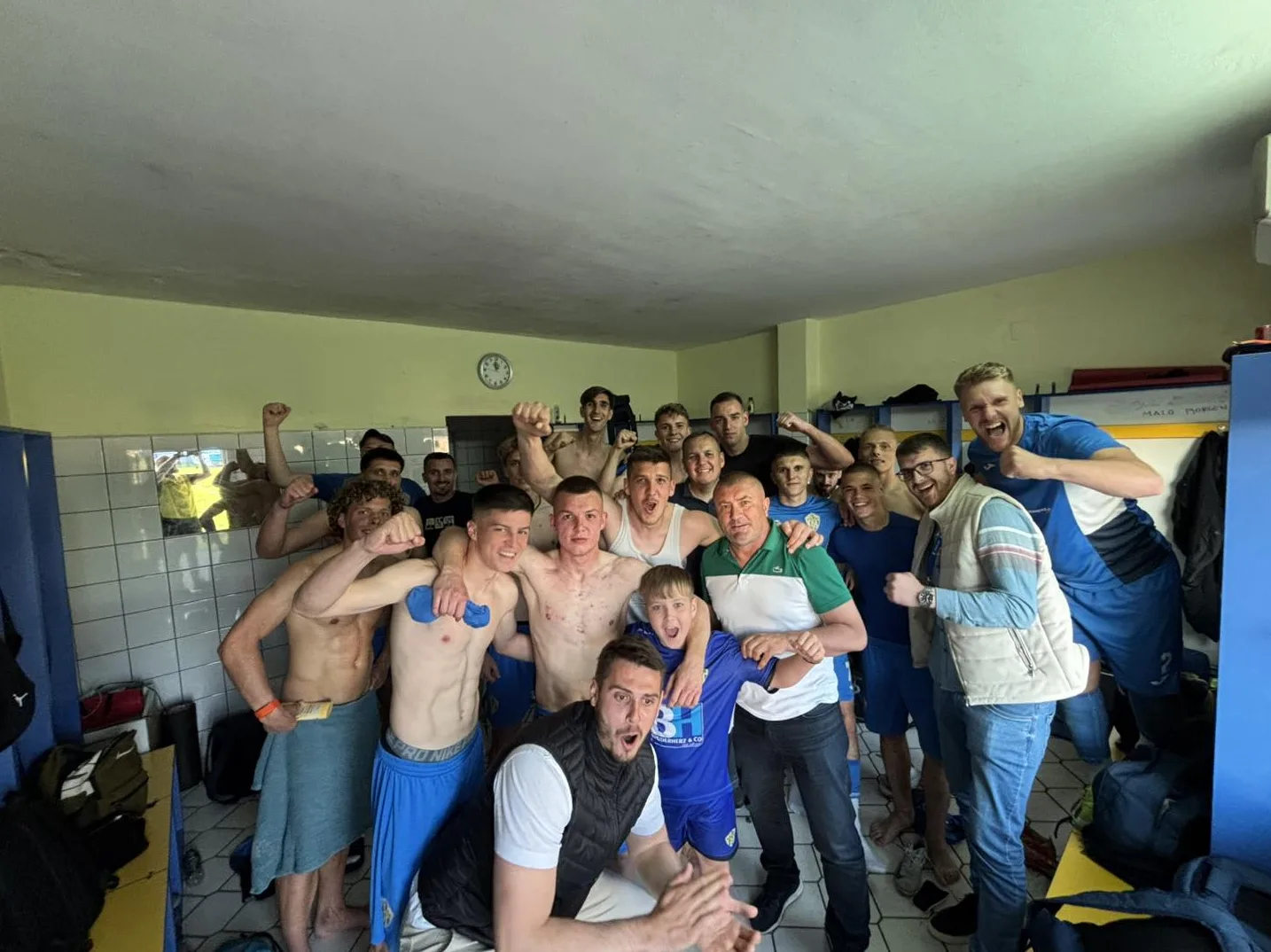 Grupa fudbalera u svlačionici podiže ruke u znak pobjede, neki su bez majica, neki nose sportske dresove.