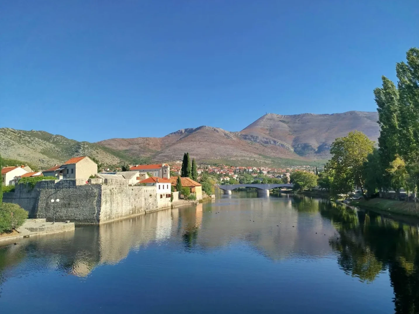 Kamene zidine Starog grada u Trebinju uz zelenu rijeku Trebišnjicu i planine u pozadini pod jasnim plavim nebom.