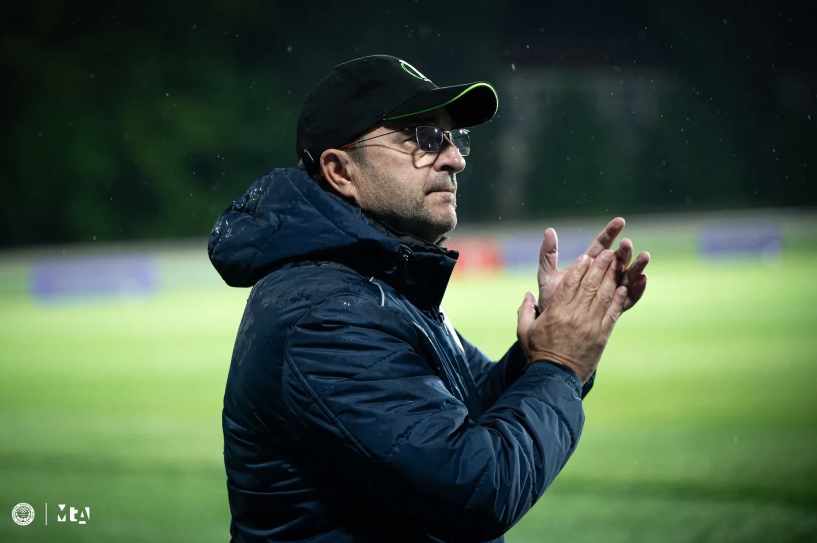 Trener u kapu i naočalama aplaudira na nogometnoj utakmici, obučen u zimsku jaknu.