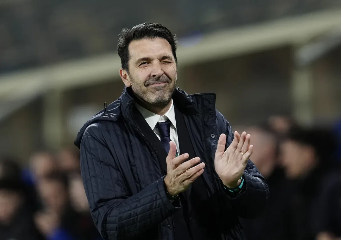 Fudbalski trener sa tamnom kosom i bradom, odjeven u tamnu sportsku jaknu preko bijele košulje i tamne kravate, plješće rukama i žmiri lijevim okom.