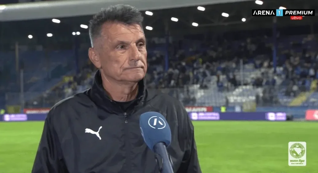 Fudbalski trener sa sijedom kosom, odjeven u crni sportski duks sa bijelim logom marke Puma, stoji na terenu stadiona i daje izjavu za medije. Mikrofon sa logom Arena Sport kanala je ispred njega. U pozadini se vide reflektori i tribine stadiona popunjene gledaocima.