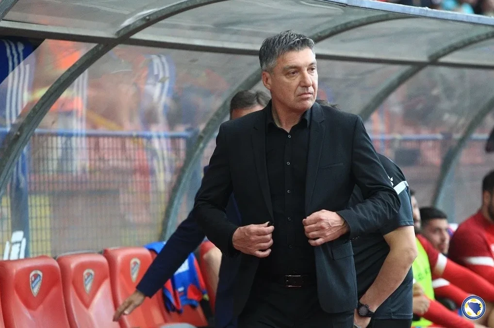 Muškarac srednjih godina u crnom odijelu i crnoj košulji stoji na stadionu, popravljajući rever odijela. U pozadini se naziru crvene stolice i drugi ljudi.