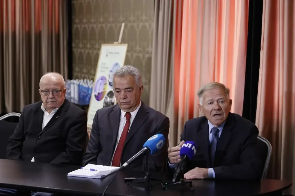 Tri muškarca sjede za crnim stolom ispred crvenkastih zastora, a ispred njih se nalaze mikrofoni s logom N1.