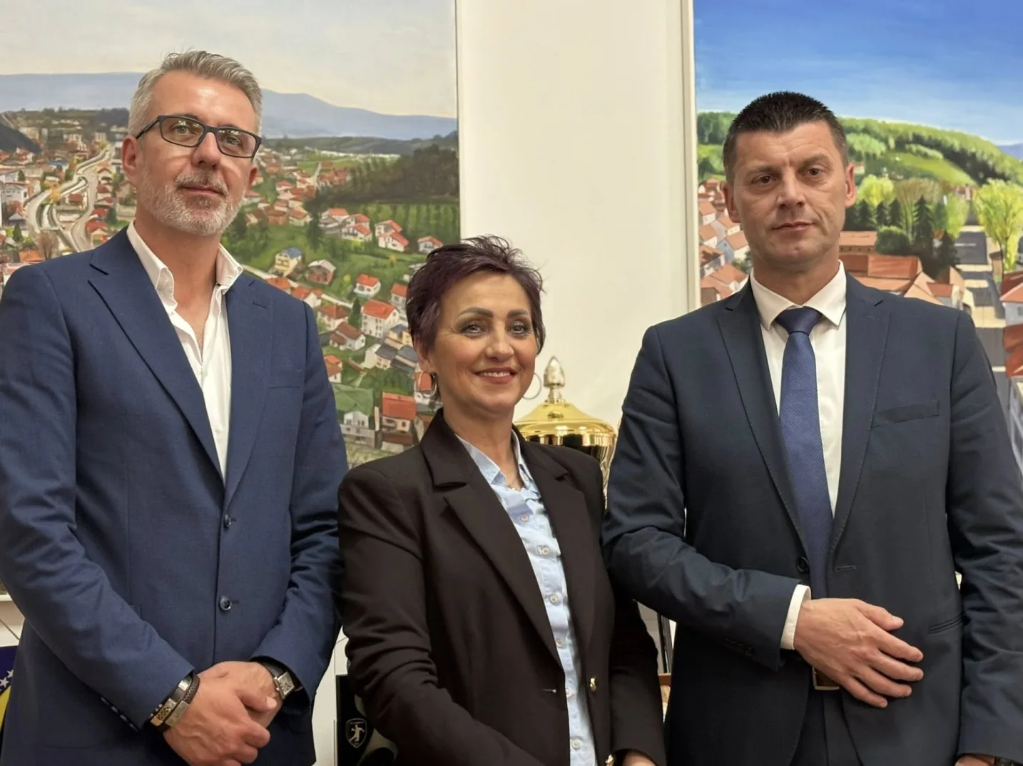 Muškarac u plavom odijelu s naočalama, žena u crnom sakou i plavoj košulji, te muškarac u tamnom odijelu s plavom kravatom stoje zajedno ispred slika koje prikazuju gradski pejzaž.