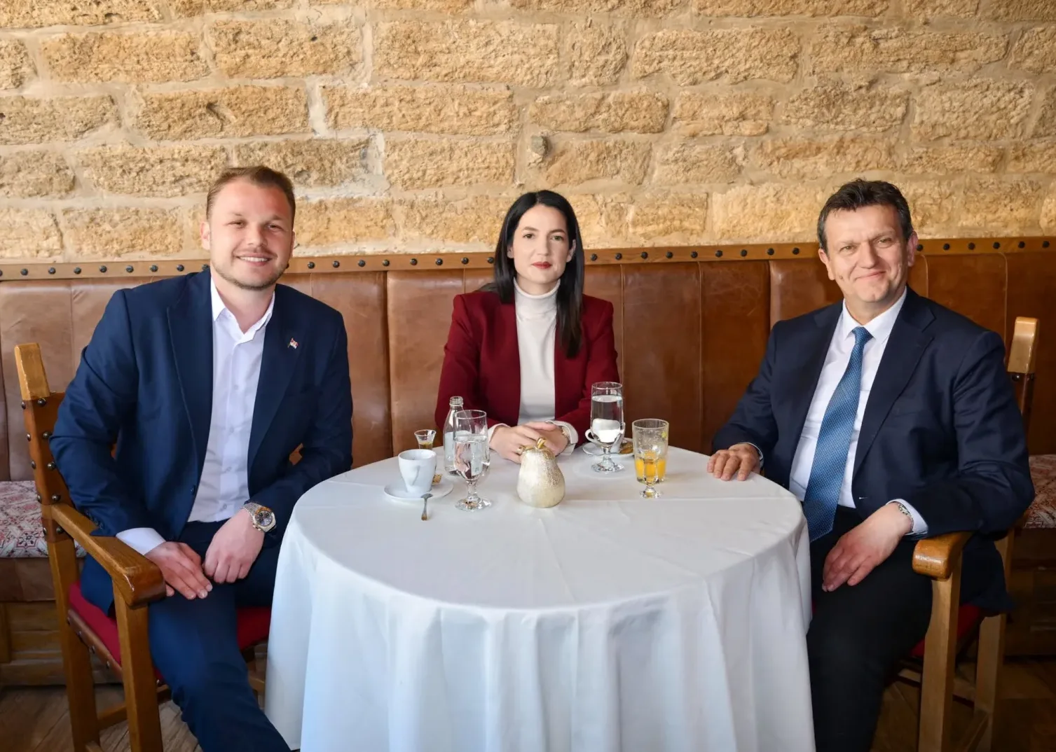 Tri osobe, dva muškarca i jedna žena, sjede za okruglim bijelim stolom u restoranu.