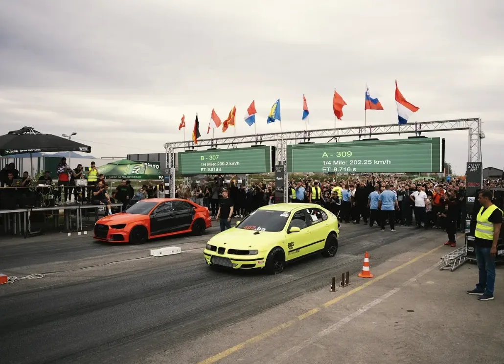 Dva automobila, narandžasto-crni i žuti, na pisti za trkanje ispred velikog broj gledalaca i semafora.