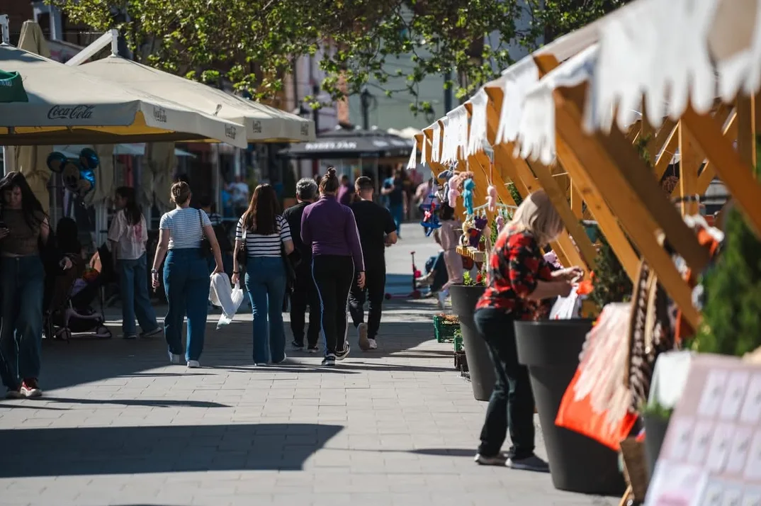 Grupa ljudi šeta popločanom ulicom pored tezgi na otvorenoj tržnici, s naglaskom na prednji dio s tezgom punom rukotvorina i ljudima koji pregledavaju robu.