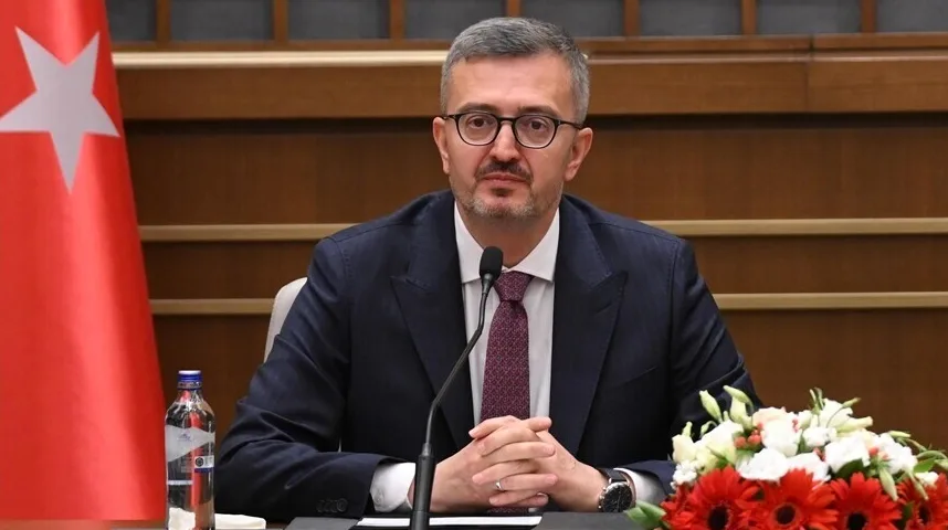 Čovjek u tamnom odijelu, bijeloj košulji i ljubičastoj kravati sa uzorkom, sjedi za stolom ispred mikrofona i cvijeća.