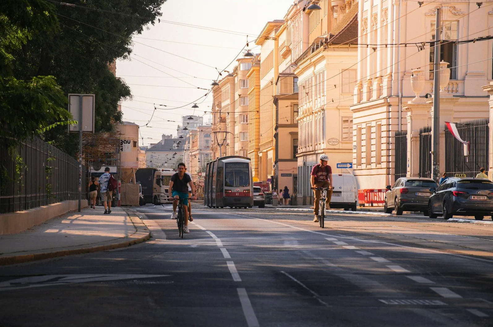 Dva biciklista voze ulicom u gradu, jedan pored drugog, dok tramvaj prolazi u pozadini. Sunčeva svjetlost pada na zgrade i ulicu.