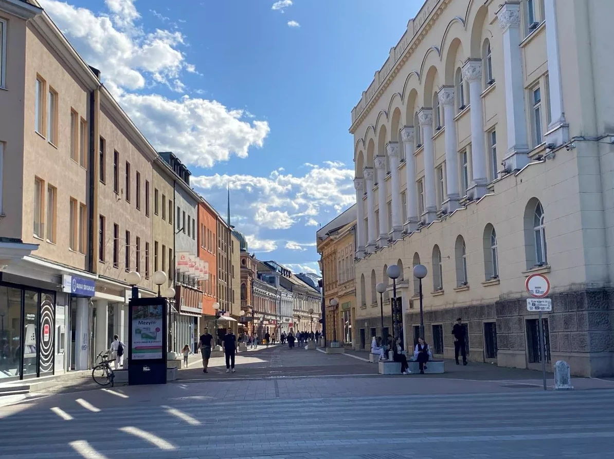 Široki pješački prelaz na suncem obasjanoj ulici sa zgradama na obje strane, punim bijelih stubova i lukova na desnoj strani, te raznobojnih fasada na lijevoj.