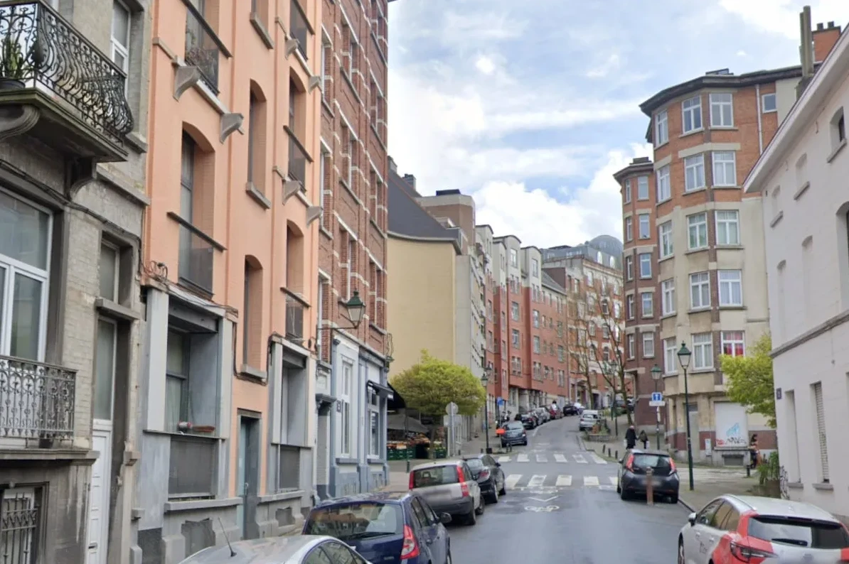 Urbani pejzaž sa stambenim zgradama različitih fasada i boja, te parkiranim automobilima duž uske ulice koja se blago uspinje.