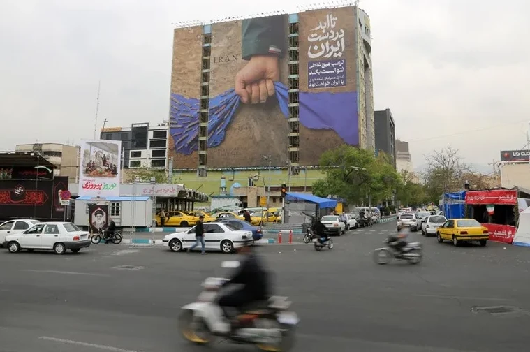Ulični pejsaž u Iranu, saobraćaj, zgrade i veliki bilbord sa rukom koja drži plavu tkaninu i natpisom "Iran"