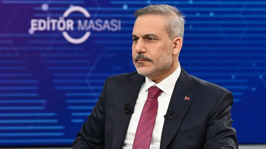 Muškarac u tamnom odijelu i crvenoj kravati sjedi u studiju s natpisom "EDİTÖR MASASI" u pozadini.