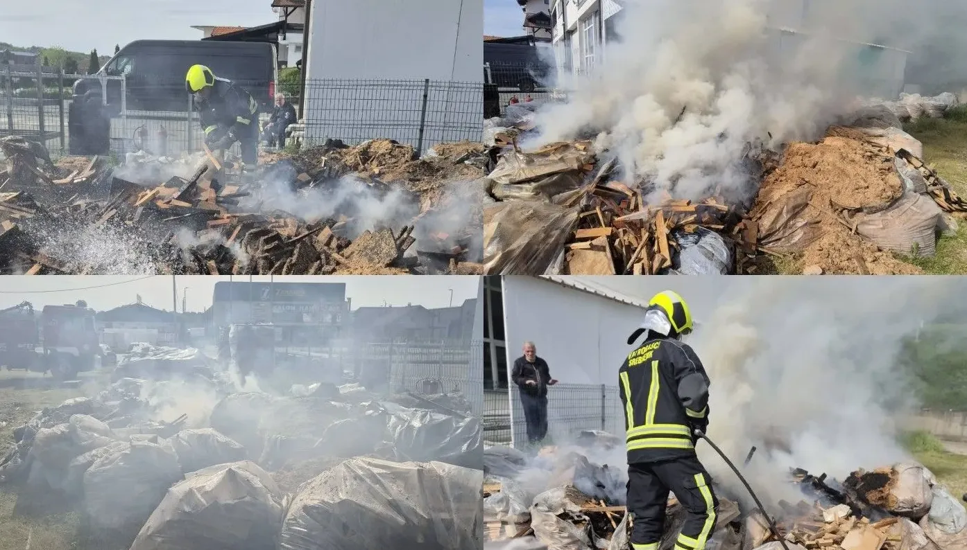 Vatrogasci u zaštitnim odijelima gase požar zapaljenog drveta i otpada, sa dosta dima.