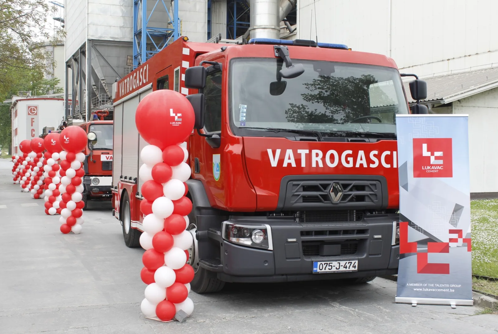 Crvena vatrogasna vozila sa natpisom "Vatrogasci", poredani redovi crvenih i bijelih balona, te baner "Lukavac Cement"